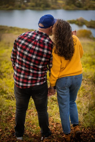 一个卷曲头发的女人吻着她的丈夫的脸颊一个穿着黄色毛衣和蓝色牛仔裤的女人 那个男人穿着格子衬衫 黑色牛仔裤和蓝色的帽子 望着秋天的河流 — 图库照片