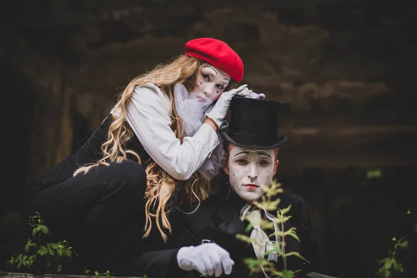 A girl in a red hat with white makeup on her face put her hands on the head of a guy in a black suit with a hat and makeup. Happy Halloween