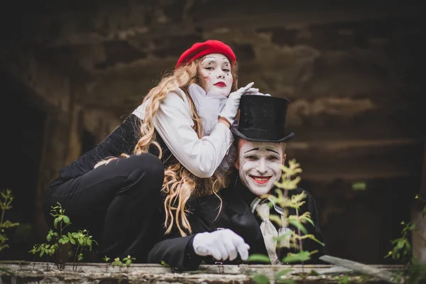 A girl in a red hat with white makeup on her face put her hands on the head of a guy in a black suit with a hat and makeup. The boy smiles terribly. Happy Halloween.