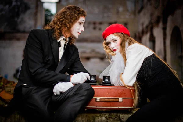 Sitting for tea with a man with makeup in a black suit and a girl in white gloves, a red hat with long hair. Halloween celebration at the ruins of the castle. Cups of tea on a suitcase.