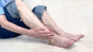 Kanepede oturan ve bacak ağrısından muzdarip bir adamın görüntüsü. Ayak bileği hastalığı, eklem iltihabı veya artrosit..