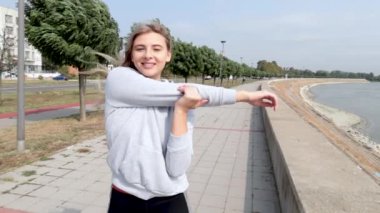 Genç, güzel, mutlu spor spor fitness kadını nehir kenarında durup kaslarını esnetiyor. Koşu ve koşuya hazırlanıyor. Bu sırada antrenman ve motivasyon için hazırlanıyor.