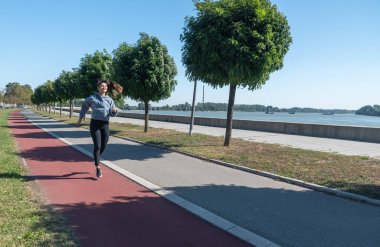 Genç, mutlu ve kendini beğenmiş koşucu kadın sağlık hayatı için nehir kenarında koşuyor ve koşuyor. Koşu yaparken motivasyon müziği dinliyor. 