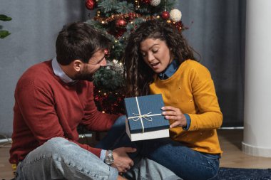 Mutlu, kaygısız genç çift Noel ağacının önünde oturmuş süslerle süslenmiş yeni yıl hediyelerini değiş tokuş ediyor ve neşeli havayı dört gözle bekliyor.