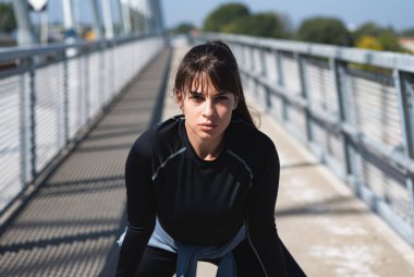 Kadın koşucu, koşusu sırasında ara veriyor. Sokakta duruyor. Kadın sporcu idmandan sonra dinleniyor, ellerini dizlerinin üstüne koyuyor. Genç terli sporcu kadın koşudan sonra dinleniyor ve koşuyor. 