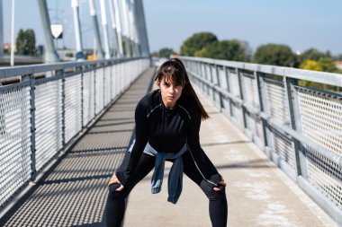 Kadın koşucu, koşusu sırasında ara veriyor. Sokakta duruyor. Kadın sporcu idmandan sonra dinleniyor, ellerini dizlerinin üstüne koyuyor. Genç terli sporcu kadın koşudan sonra dinleniyor ve koşuyor. 