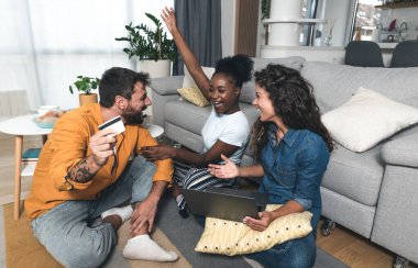 Üç heyecanlı arkadaş internetten tatil bileti alıyorlar. Kredi kartı ve dizüstü bilgisayarla evdeki oturma odasında oturuyorlar. Bilgisayarla online alışveriş yapan bir grup insan.