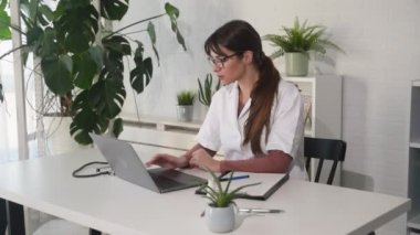 Beyaz önlüklü ve gözlüklü kadın doktor hastanede dizüstü bilgisayarında daktilo kullanıyor. Klinikte çalışan bir kadın doktor. Sağlık, teknoloji insanları ve tıp konsepti. 