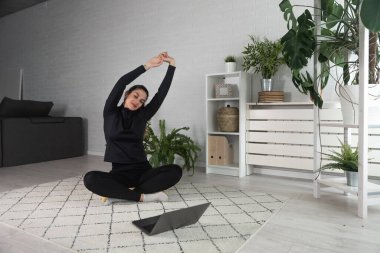 Nilüfer pozisyonunda oturan mutlu ve çekici genç bir kadın antrenörle internetten yoga kursuna başlıyor ya da uzaktan antrenör olarak Webinar seansı yapıyor..