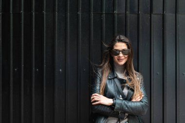 Deri ceketli, güneş gözlüklü, siyah duvarda duran güzel bir kadının portresi. Sevimli kadın pozitifliği ve ellerini çapraz yapıp saçlarını sallarken dışarıda poz vermesi..