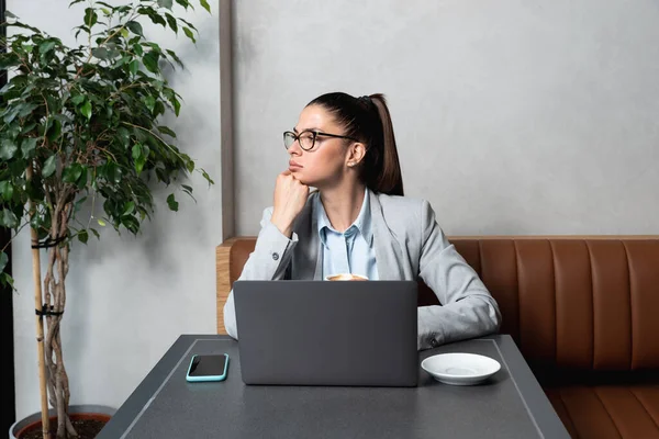 Young business woman sitting in cafeteria drinking coffee and working on her laptop waiting for waiter and her food to arrive. Female businessperson on lunch break wait to order her meal.