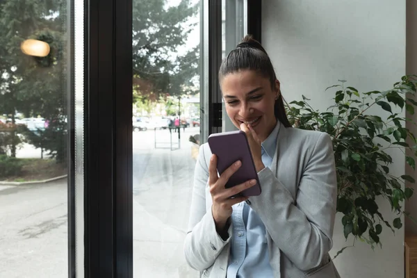 Young business woman in casual suit standing near window in the restaurant holding her smartphone waiting to waiter pack her lunch to go in the box. Freelancer expat waiting take away coffee or food.