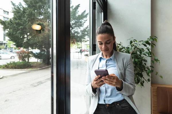 Young business woman in casual suit standing near window in the restaurant holding her smartphone waiting to waiter pack her lunch to go in the box. Freelancer expat waiting take away coffee or food.