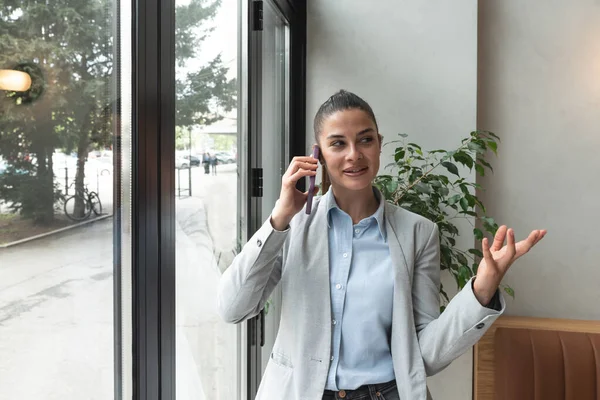 Young business woman in casual suit standing near window in the restaurant holding her smartphone waiting to waiter pack her lunch to go in the box. Freelancer expat waiting take away coffee or food.