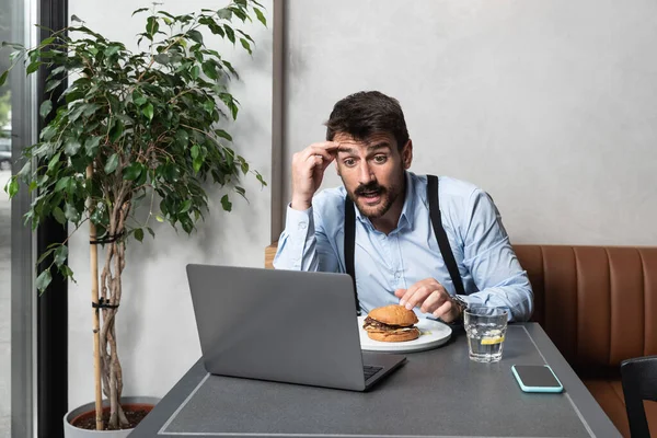 Young successful business man freelancer with good mood using laptop computer for distance work during lunch break in cafe bar, attractive male sitting in cafeteria coffee shop eating burger and work.