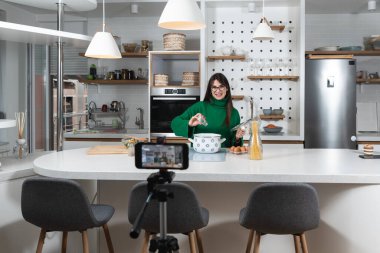 Genç vlogger kadın, apartmanında yemek pişirmekten ve internet için internet yaratıcısı olarak akıllı telefonlu video bloğu yapmaktan bahsediyor. Kadın nüfuzlu biri yemek yapıyor..