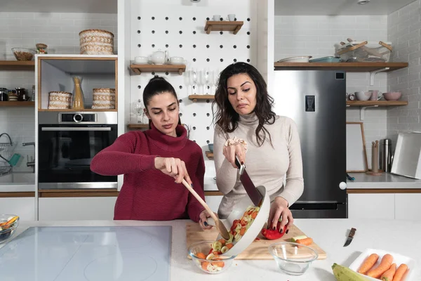 Two young happy college student roommates or business women cooking food together at their apartment. Females having fun together preparing meal in their modern kitchen at cozy home.