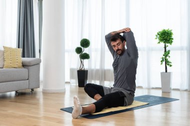 Happy attractive young business man in active sport wear sitting on floor stretching muscles at his apartment after or before the work, training yoga class. Home workout as stress and pain relief.