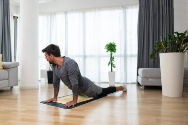 Happy attractive young business man in active sport wear sitting on floor stretching muscles at his apartment after or before the work, training yoga class. Home workout as stress and pain relief.