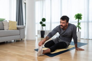 Happy attractive young business man in active sport wear sitting on floor stretching muscles at his apartment after or before the work, training yoga class. Home workout as stress and pain relief.