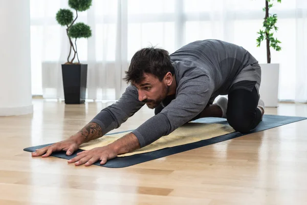 Happy attractive young business man in active sport wear sitting on floor stretching muscles at his apartment after or before the work, training yoga class. Home workout as stress and pain relief.