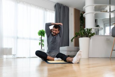 Happy attractive young business man in active sport wear sitting on floor stretching muscles at his apartment after or before the work, training yoga class. Home workout as stress and pain relief.
