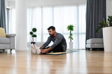 Happy attractive young business man in active sport wear sitting on floor stretching muscles at his apartment after or before the work, training yoga class. Home workout as stress and pain relief.
