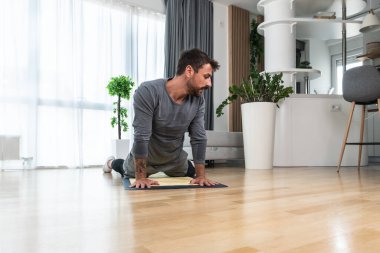 Happy attractive young business man in active sport wear sitting on floor stretching muscles at his apartment after or before the work, training yoga class. Home workout as stress and pain relief.