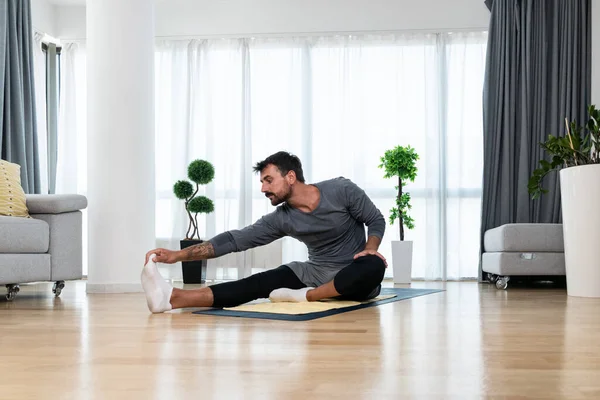 Happy attractive young business man in active sport wear sitting on floor stretching muscles at his apartment after or before the work, training yoga class. Home workout as stress and pain relief.