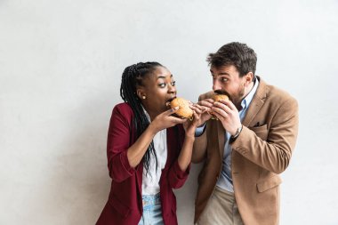 Two young business people or smart casual white man and African American woman holding and eating fat tasty American hamburger or burger while they making funny faces and goofing. Happy people eating 