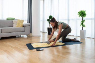 Happy attractive young business woman in active sport wear sitting on stretching muscles at her apartment after or before the work, training yoga class. Home workout as stress and pain relief.