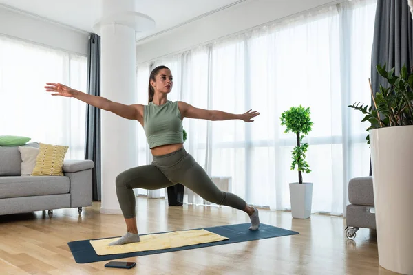 Happy attractive young business woman in active sport wear sitting on stretching muscles at her apartment after or before the work, training yoga class. Home workout as stress and pain relief.