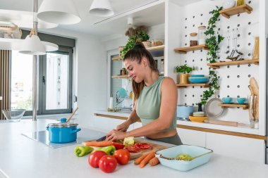 Young sporty woman cutting and cooking fresh natural vegetables at kitchen and cooking for healthy lunch. Self care yoga nutritionist girl having healthy meal after workout.