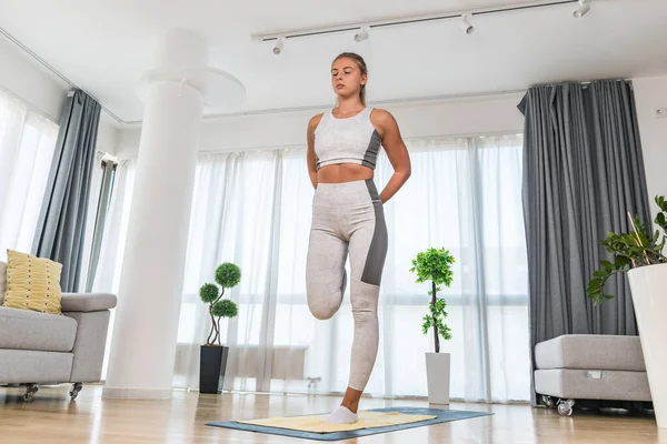 Happy attractive young business woman in active sport wear sitting on stretching muscles at her apartment after or before the work, training yoga class. Home workout as stress and pain relief.