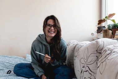 Young lady feeling happy about getting pregnant that she find from pregnancy test she is holding while sitting on bed. Woman with happiness facial expression future mother positive surprise from test.