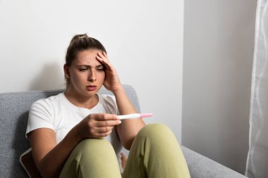 Young nervous woman is sitting at home holding a pregnancy test and is impatiently waiting for the test result. Maybe pregnant maybe not concept with anxious inpatient female on sofa in her apartment.