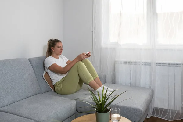 Young nervous woman is sitting at home holding a pregnancy test and is impatiently waiting for the test result. Maybe pregnant maybe not concept with anxious inpatient female on sofa in her apartment.