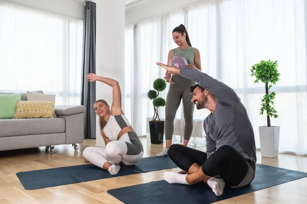 Young woman fitness and yoga instructor helping couple in their home for workout exercises. Female coach working with clients in their apartment. Husband and wife paying physical therapist for sport.