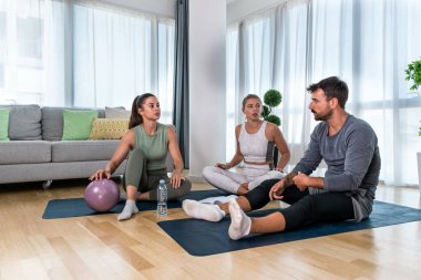 Young woman fitness and yoga instructor helping couple in their home for workout exercises. Female coach working with clients in their apartment. Husband and wife paying physical therapist for sport.