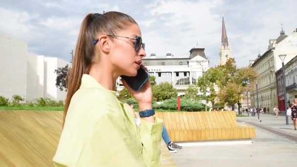 时髦的黑发女人拿着智能手机 在讲电话 穿着漂亮的衣服 戴着太阳镜在街上走着 时尚春夏户外 谈生意的女商人 — 图库视频影像