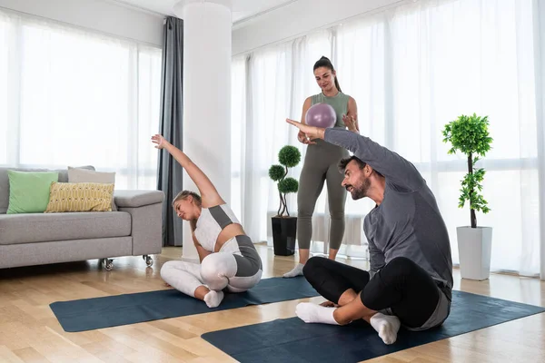 Young woman fitness and yoga instructor helping couple in their home for workout exercises. Female coach working with clients in their apartment. Husband and wife paying physical therapist for sport.