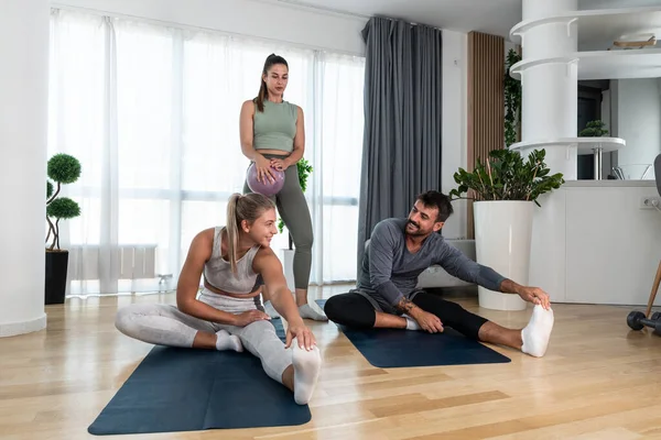 Young woman fitness and yoga instructor helping couple in their home for workout exercises. Female coach working with clients in their apartment. Husband and wife paying physical therapist for sport.