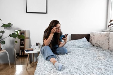 Makyaj aynası yerine akıllı telefon kamerası kullanan cımbızla kaş koparan genç bir kadın. Yüzündeki kılları keserken kızın yüzü. Kaş güzelliği düzeltir.
