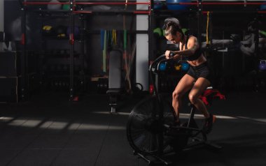 Genç, terli, güçlü sporcu bir kadın spor salonunda bisiklet sürüyor ya da koşuyor. Güç ve kondisyon için yoğun kardiyo antrenmanı için.