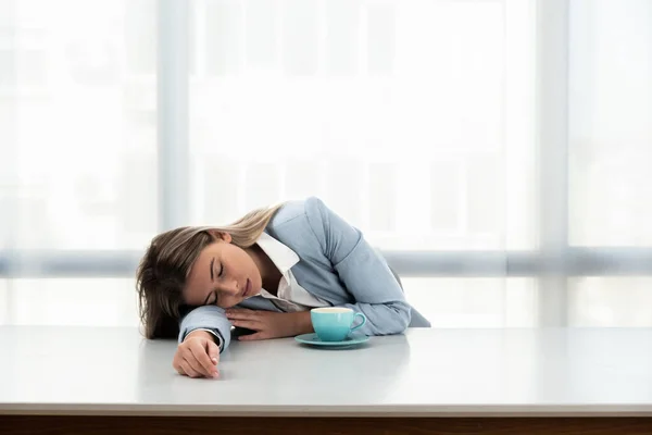 Young tired business woman sleeping at the empty office desk with one cup of coffee feeling overworked. Businessperson at work in the morning.