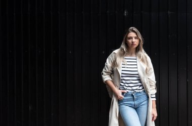 Kareli kot pantolon ve bej renkli trençkot giymiş çekici bir kadının portresi. Dışarıya bakıyor ve poz veriyor. Havalı bir kadın, rahat ve şık bir kıyafetle dışarıda poz veriyor. 