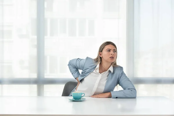 Young business woman sitting in the office in formal wear at empty desk with cup of coffee have a back pain from long stressful sitting at the work chair. Discus Hernia concept.