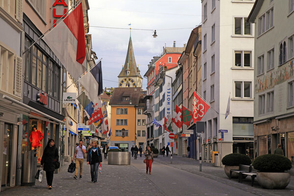 Zurich, Switzerland - May 04, 2013: Cobbled pedestrian street Rennweg with boutique shops, flags on old buildings and St. Peter church clock tower in Zurich, Switzerland. Zurich is international capital of finance and center of banking industry.