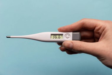 Mavi arka planda termometreyi tutan kadın. Termometre normal sıcaklığı gösteriyor.