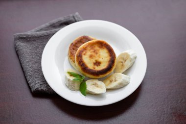 Two cottage cheese pancakes with sliced banana in a white plate. Traditional breakfast or lunch. Traditional Ukrainian breakfast.